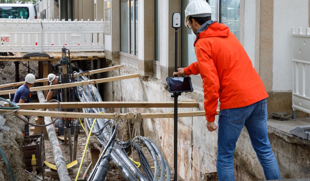 Image shows guy performing a survey using measuring equipment in a large ditch by a bulding with utility pipes
